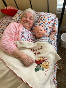 My mom laying in a twin size bed with her baby doll in her arms, covered with a soft blanket.